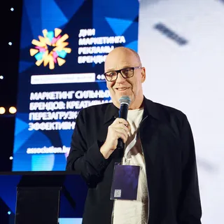 Спикер в очках выступает с микрофоном на сцене конференции «Дни маркетинга, рекламы и брендинга» на фоне брендированного экрана