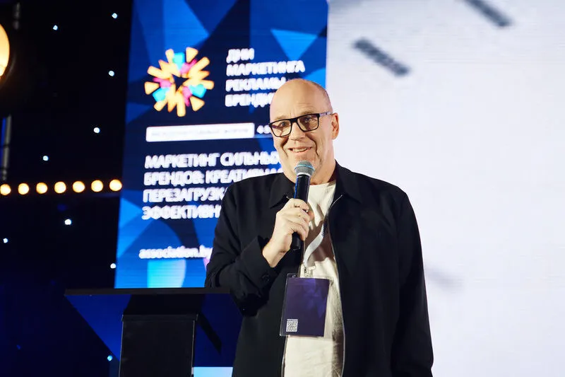 Спикер в очках выступает с микрофоном на сцене конференции «Дни маркетинга, рекламы и брендинга» на фоне брендированного экрана