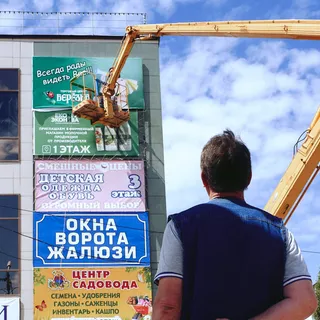 Мужчина смотрит на фасад здания, увешанный рекламными баннерами разных магазинов, рядом с ним — жёлтый автогидроподъёмник для их установки