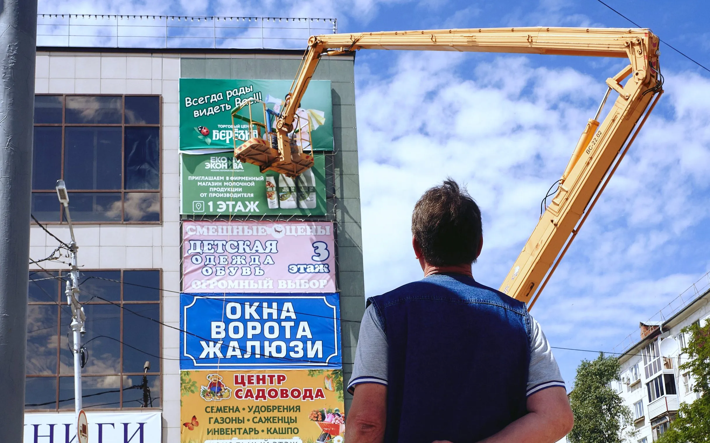 Мужчина смотрит на фасад здания, увешанный рекламными баннерами разных магазинов, рядом с ним — жёлтый автогидроподъёмник для их установки