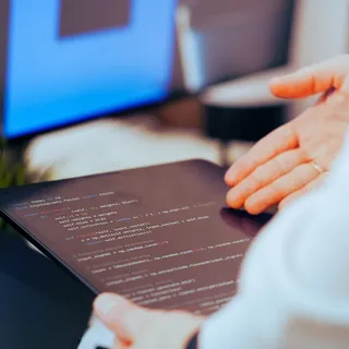 Крупным планом планшет с фрагментом кода на Python (нейросеть, работа с векторами) в руках у человека, на фоне размытый монитор с синим экраном