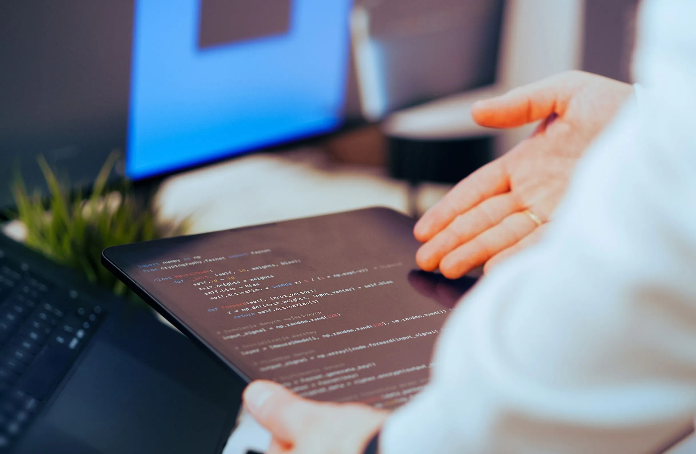 Крупным планом планшет с фрагментом кода на Python (нейросеть, работа с векторами) в руках у человека, на фоне размытый монитор с синим экраном