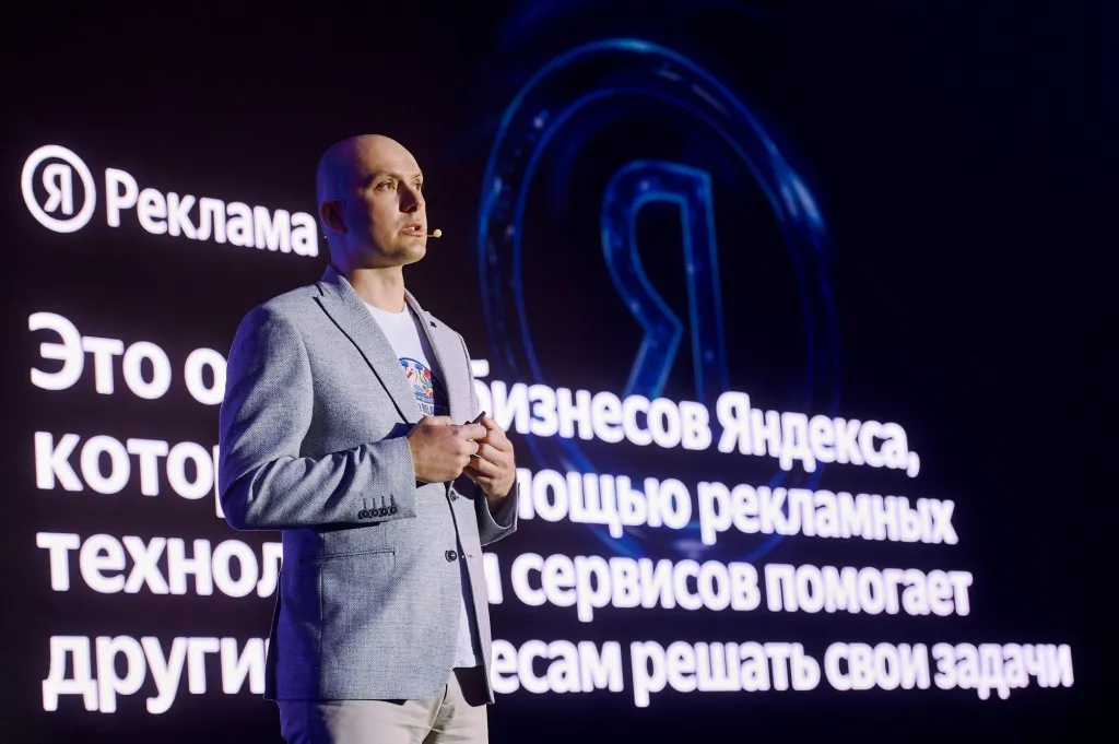 Спикер в сером пиджаке выступает на сцене конференции Яндекс Рекламы на фоне экрана с логотипом «Я Реклама»