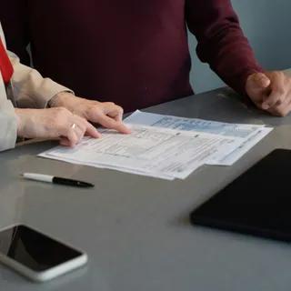 Двое за столом переговоров изучают документ с таблицей, рядом лежат смартфон, планшет и ручка